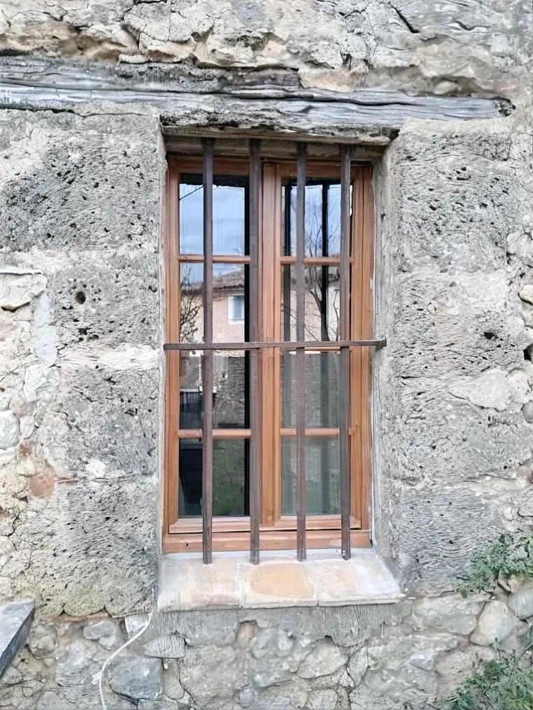 Façade extérieure en pierre naturelle avec fenêtre à double vantaux en bois équipée de barreaux de sécurité métalliques
