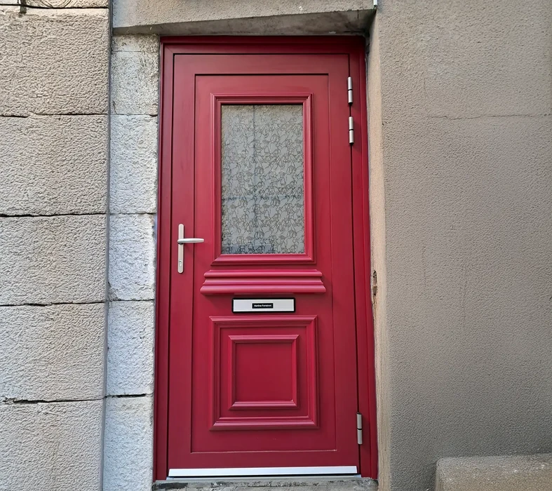 Entrée extérieure d'une habitation avec porte d'entrée rouge bordeaux en PVC ou bois, équipée d'un vitrage décoratif texturé dans la partie haute et d'un panneau décoratif en bas