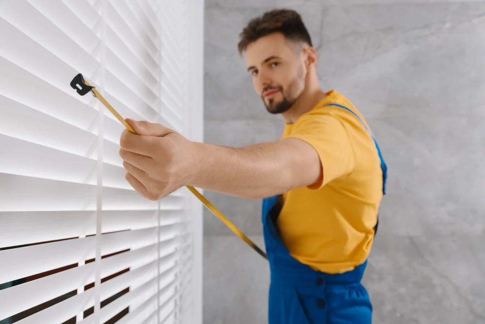 Un homme en salopette bleue et t-shirt jaune nettoie des stores vénitiens blancs avec un plumeau à manche jaune dans un intérieur moderne aux murs blancs