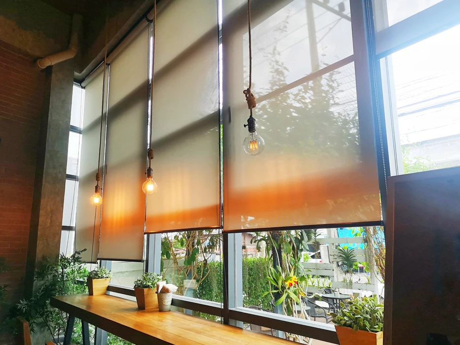 Intérieur moderne avec grandes baies vitrées, stores enrouleurs dégradé orange installés, ampoules suspendues style industriel, plantes vertes en pots sur rebord de fenêtre