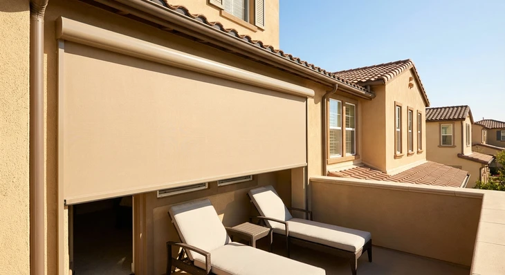 Balcon résidentiel étroit d'une maison individuelle avec un store extérieur motorisé beige déployé, offrant une ombre complète sur l'espace du balcon et ses chaises longues, par une journée ensoleillée. Le store doit être clairement visible en position déployée protégeant l'espace réduit du balcon. Architecture résidentielle classique en arrière-plan avec ciel bleu dégagé.