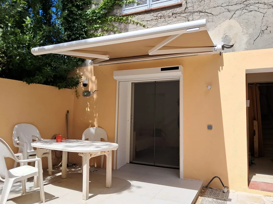 Terrasse extérieure d'une maison individuelle avec un store extérieur toile déployé, offrant une ombre généreuse. Le store est le point focal de l'image, clairement visible avec ses structures de fixation. La terrasse accueille du mobilier de jardin confortable, et on voit l'arrière de la maison en arrière-plan. L'ambiance est lumineuse et chaleureuse, mettant en valeur le confort et la protection solaire apportés par le store. La photo est prise en conditions réelles, en bon temps, montrant le résultat d'une installation professionnelle.