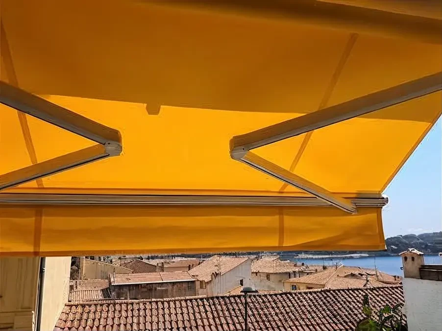 Terrasse moderne d'une maison résidentielle de l'Est-Var, vue de trois-quarts, avec un store extérieur de couleur gris ou taupe déployé couvrant la zone de détente. Le store banne affiche clairement ses bras articulés métalliques et son toile tendue en configuration ouverte, créant une zone d'ombre bien définie. La terrasse dallée accueille un coin lounge avec fauteuils modernes et une table basse. Un petit jardin paysager avec massifs de plantes méditerranéennes encadre la scène. La photo est prise en plein jour ensoleillé, mettant en relief le contraste lumière/ombre et l'intégration élégante du store sur la façade de la maison. Atmosphère résidentielle professionnelle et accueillante.